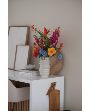 En vit räfflad vas med Artificiell Blomma Kattmynta 75 cm rosa står på ett vitt skrivbord med böcker, inramad konst, en träbricka och en skärbräda, bredvid en beige stol i ett modernt minimalistiskt rum.