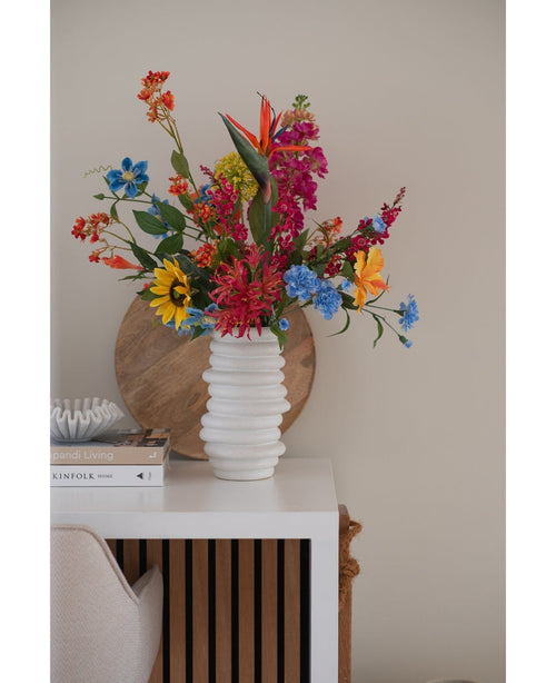 En vit texturerad vas rymmer en konstgjord blomma kattmynta 75 cm rosa bukett på ett vitt bord bredvid staplade böcker och dekor, med en rund träbricka lutad mot den beiga väggen i bakgrunden.