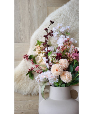 Den konstgjorda blomman Jilly 70 cm rosa konstgjord blomma står i en beige vas på ett trägolv bredvid en mjuk elfenbensfärgad fårskinnsmatta.