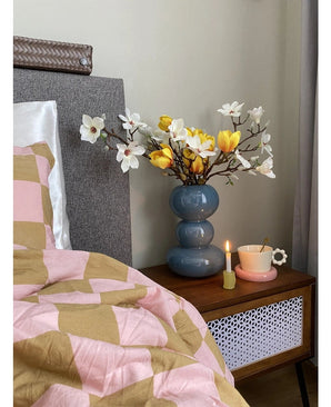 Ett sängbord med en blå vas innehållande den konstgjorda blomman Magnolia 50 cm gul/orange, en rosa tekopp på ett prästkrageformat fat och ett brinnande ljus står bredvid en säng täckt med en rosa och brun filt med geometriska mönster.