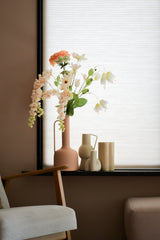 En pastellfärgad vardagsrumshörna med en trästol, en mjuk fotpall och en fönsterbräda med Artificiell Blomma Fritillary 94 cm vit konstgjord blomma i en rosa vas bredvid minimalistisk keramisk dekor mot vita persienner.