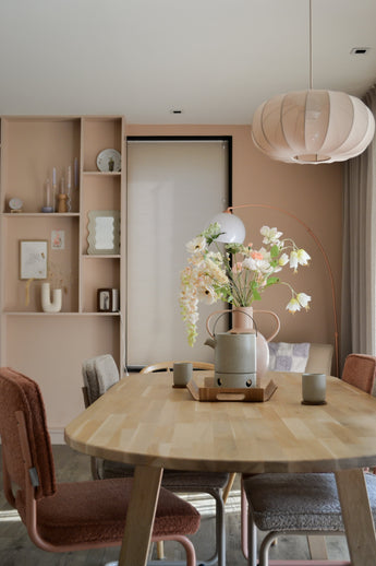 En modern matsal med ett ljust träbord, fyra stolar med kuddar, konstgjord blomfritillary 94 cm vit i vas, keramiska koppar, pastellrosa väggar, inbyggda hyllor med dekor och en stor rund hängande lampa ovanför ditt huvud.