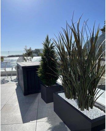 Kunstgras Bordeaux UV 120cm in rechthoekige plantenbakken met witte stenen siert een modern buitenterras tegen een achtergrond van blauwe lucht en bomen in de verte.