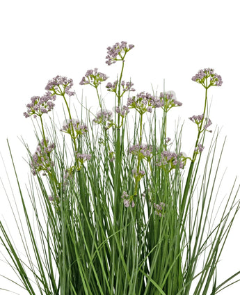 De Kunstgras Prei Bloem Ui Graspot 60cm heeft hoge, dunne groene bladeren en trossen kleine, delicate lichtpaarse bloemen op lange stelen. Het arrangement is brandvertragend en afgebeeld tegen een witte achtergrond.