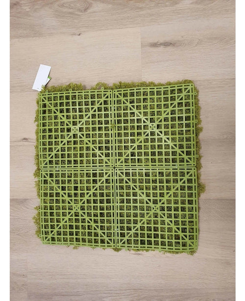 Artificiell Haag Nordic Moss 50x50 cm UV är en grön konstgräsmatta med en synlig plastruta på baksidan, idealisk för lyxiga interiörer eller för att skapa en grön vägg. Den vilar på ett ljust trägolv med en liten vit etikett i ena hörnet.