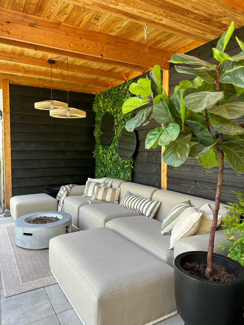 En mysig uteservering med en beige soffa, randiga kuddar, en rund eldstad, en stor planta i krukor och en träpergola. Väggen är dekorerad med grönska med Artificiell Haag Volta 100x100 cm UV för extra stil.