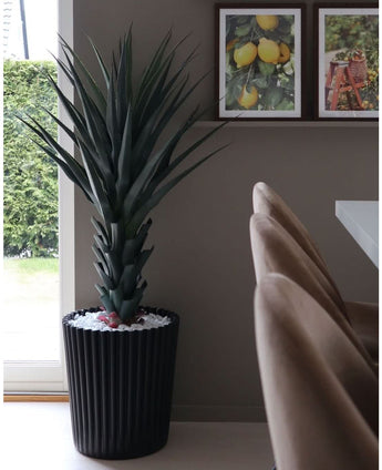 De Kunstplant Agave Pandora UV 120 cm, een hoge stekelige groene plant in een zwarte geribbelde pot met witte stenen, staat bij het raam. Daarachter hangen drie ingelijste fruitprints op een wandplank boven beige stoelen en een witte tafel.