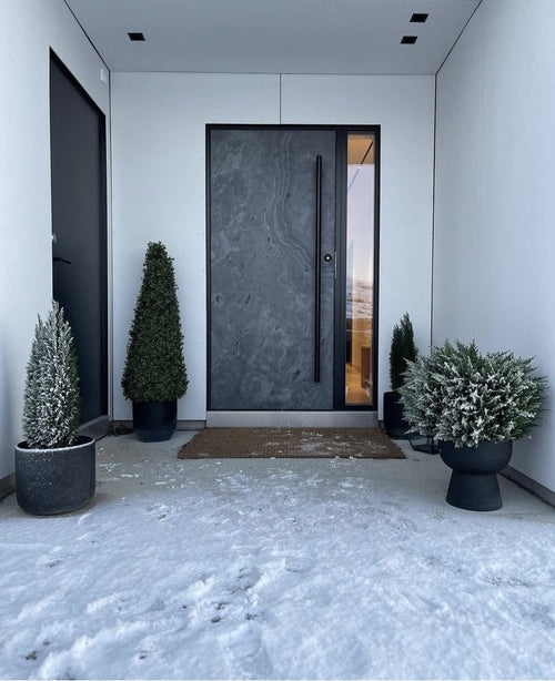 En modern mörk ytterdörr ramas in av vita väggar och flankeras av Artificiell växt buxbom Pyramid UV 90 cm, som ger UV-skydd. Lätt snö täcker marken, medan dessa snygga trädgårdsväxter skapar en snygg, minimalistisk entré.