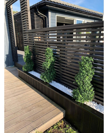 Tre konstgjorda buxbomsspiraler i romersk stil UV 90 cm topiary buskar står i en smal bädd av vita stenar, mot ett modernt svart ribbad insynsskydd bredvid en trä uteplats och ett modernt hus.