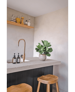 En modern bänkskiva med mässingskran, två tvålautomater, artificiell tobaksväxt Fiddle Leaf UV 45 cm krukväxt och två träpallar. En flytande hylla med burkar och flaskor är monterad på en ljus vägg.
