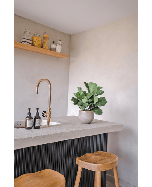 En modern bänkskiva med mässingskran, två tvålautomater, artificiell tobaksväxt Fiddle Leaf UV 45 cm krukväxt och två träpallar. En flytande hylla med burkar och flaskor är monterad på en ljus vägg.