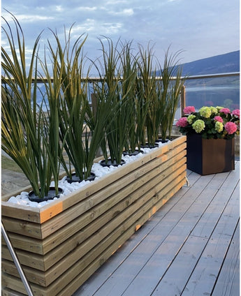 En träkruka med konstgräsgrön UV 120cm och vita stenar står på en balkong med glasräcke; i närheten, en svart fyrkantig kruka med blommande rosa, gröna och vita hortensior med utsikt över den pittoreska utsikten.