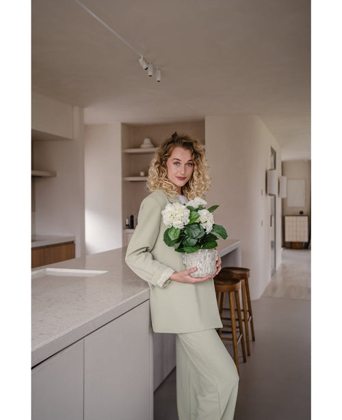 En kvinna med lockigt blont hår, klädd i en ljusgrön outfit, står i ett modernt kök med en artificiell hortensia Vit UV 35 cm växt i händerna – med krämvita blommor – och ler mot kameran.
