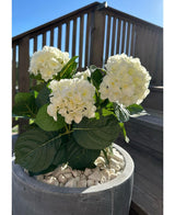 Den konstgjorda hortensia White UV 35 cm-växten, med vita blommor och gröna blad, står ute på vita småsten en solig dag, med uteplatsräcken i trä och en trappa i bakgrunden.