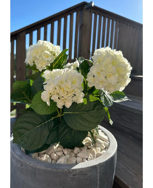 Den konstgjorda hortensia White UV 35 cm-växten, med vita blommor och gröna blad, står ute på vita småsten en solig dag, med uteplatsräcken i trä och en trappa i bakgrunden.