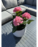 Den konstgjorda växten Hortensia Pink UV 35 cm, en vit kruka med levande rosa hortensiablommor, står på ett vävt grått utebord med en grå soffa med kuddar i bakgrunden, upplyst av solljus och mjuka skuggor.