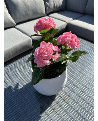 Den konstgjorda växten Hortensia Pink UV 35 cm, en vit kruka med levande rosa hortensiablommor, står på ett vävt grått utebord med en grå soffa med kuddar i bakgrunden, upplyst av solljus och mjuka skuggor.