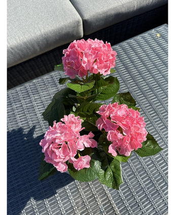 Den konstgjorda växten Hortensia Rosa UV 35 cm, med tre rosa hortensiablommor och gröna blad, står på ett vävt grått utebord, med en grå soffa med kuddar i bakgrunden.