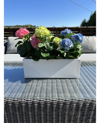 Den konstgjorda växten Hydrangea Pink UV 35 cm, en rektangulär vit plantering med rosa hortensior, placeras ute på ett flätat bord med glasskiva, gråa kuddar och en blå himmel i bakgrunden.