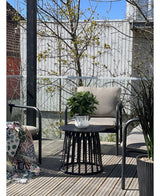 En modern terrass med två stolar med kuddar, ett runt svart bord med växten Artificiell Laurel UV d33 cm som mittpunkt och en mönstrad filt på en stol. Träd och ett trästaket syns i bakgrunden under en klar himmel.