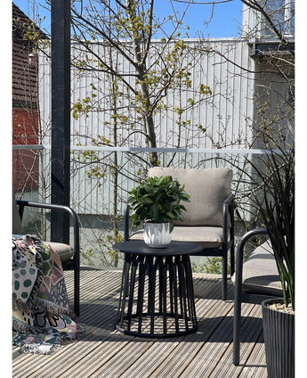 En modern terrass med två stolar med kuddar, ett runt svart bord med växten Artificiell Laurel UV d33 cm som mittpunkt och en mönstrad filt på en stol. Träd och ett trästaket syns i bakgrunden under en klar himmel.
