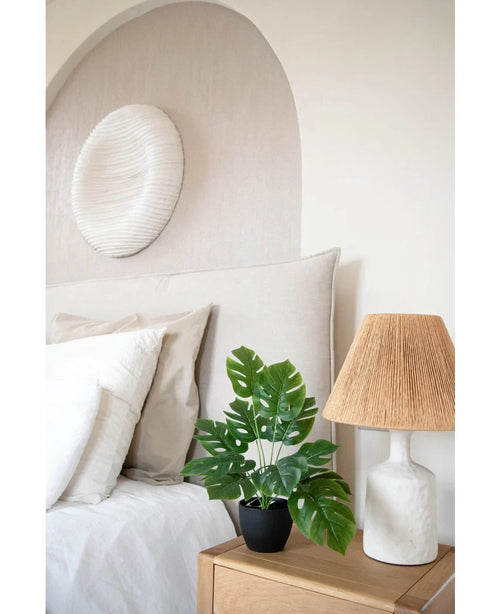 Ett modernt sovrum i neutrala toner har en vit säng med beige och vita kuddar, ett sängbord i trä med en keramisk lampa och en vävd skärm, abstrakt väggkonst ovanför sängen och Art Monstera 40 cm i en svart dekorativ kruka som tillför grönska.