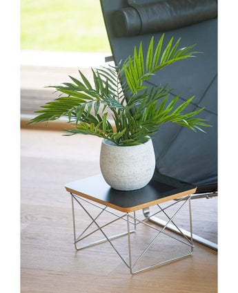 En konstgjord palmsalong UV 60 cm står i en kruka på ett litet modernt sidobord med svart skiva och metalltrådsben, bredvid en svart stol på ett ljust trägolv.
