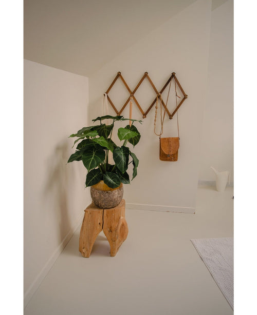 En minimalistisk rumshörna blir en grön oas med Artificiell växt Syngonium UV 58 cm på en pall i naturträ, bredvid en brun läderväska på ett geometriskt väggställ och en vit kanna, allt mot benvita väggar och golv.