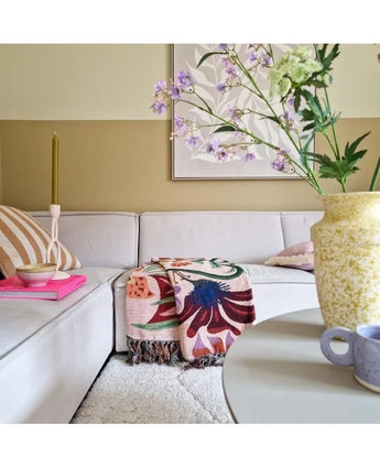 Een gezellige woonkamer met een lichtbeige sofa, een kleurrijke deken met bloemen en kussens met een patroon. Op een ronde tafel staan een gele vaas met Kunsttak Daphne Spray 108 cm paars en een klein blauw kopje. Aan de neutraal getinte muur hangt kunst.