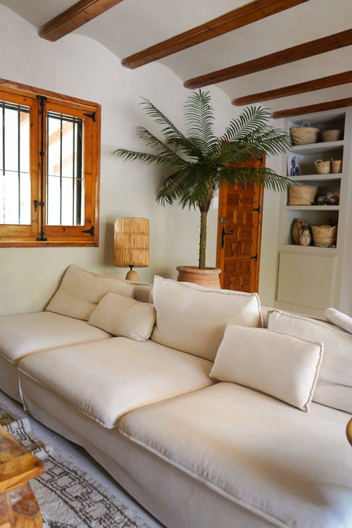 Ett mysigt vardagsrum med en stor krämfärgad soffa, Artificiell Palm Phoenix De Luxe 210 cm i hörnet, en trälampa, inbyggda hyllor med korgar, takbjälkar i trä och ett fönster med träramar.