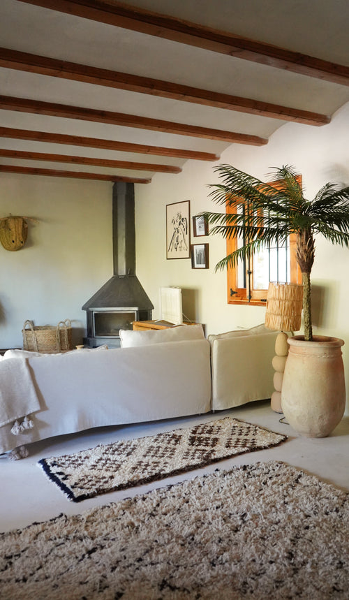 Ett mysigt vardagsrum har en vit soffa, mönstrad matta, braskamin, inramad konst och träbjälkar. Solljus strömmar in från ett fönster bredvid den frodiga Artificiella Palm Phoenix De Luxe 210 cm, vilket ger utrymmet liv.