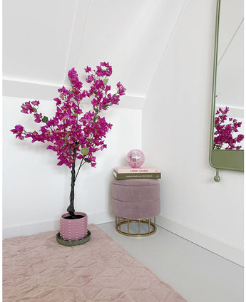 En modern, minimal rumshörna med en konstgjord bougainvillea 125 cm Lila växt i kruka, en rund rosa fotpall med guldben, staplade böcker, en rosa kristallkula, en väggspegel och en mjuk rosa matta på ett ljust golv.