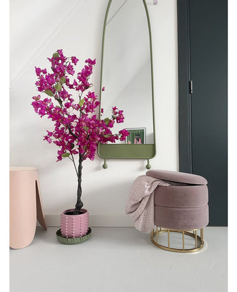 En modern inredning med Artificiell Bougainvillea 125 cm Lila i en dekorativ kruka, en hög spegel med grön ram, en rosa fotpall med guldbotten och en rosa filt och en mörk dörr mot en krispig vit vägg.