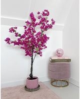 Artificiell Bougainvillea 125 cm Lila med livfulla lila blommor står bredvid en rosa sammetspall med böcker staplade ovanpå och en rosa glaskula i ett minimalt, modernt rum med vita väggar.