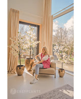 Een vrouw lacht op een beige fauteuil bij grote ramen met beige gordijnen, omringd door drie Kunstplant Bougainvillea 150 cm wit in een lichte, moderne leefruimte vol natuurlijk licht.