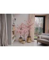 Een Kunstplant Bougainvillea 180 cm roze staat in een gouden plantenbak bij een raam in een moderne woonkamer, naast een boekenplank en beige gordijn, met rechts een grijze fauteuil zichtbaar.