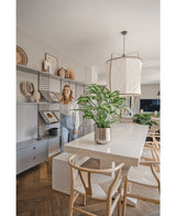 Een vrouw glimlacht in een lichte, moderne eetkamer met houten stoelen, een witte tafel met de Kunstplant Dieffenbachia 75 cm als middelpunt en open planken versierd met boeken en kunst.