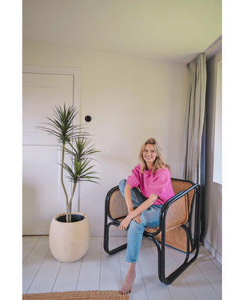 En kvinna i en knallrosa topp och blå jeans sitter barfota på en modern svart och flätad stol bredvid den konstgjorda växten Dracaena Fragnans 150 cm i ett ljust, minimalistiskt rum med vita väggar och golv.