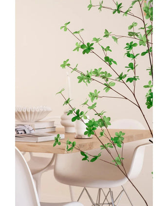 Een moderne eethoek met witte stoelen en een lichthouten tafel, gedecoreerd met boeken, kaarsen en een witte vaas, wordt op de voorgrond omlijst door de Kunstplant Enkianthus 150 cm bladgroene plant.