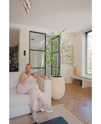 En kvinna i ljusa loungekläder sitter i en beige soffa och ler med en kopp kaffe i ett modernt vardagsrum med trägolv, fönster från golv till tak och Artificiell Enkianthus 180 cm som en stor krukväxt.