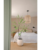 Ett minimalistiskt sovrum med växten Artificiell Enkianthus 180 cm som stor krukväxt, en bädd med vitt strö, en vävd hänglampa, beiga väggar och en naturfibermatta för en enkel och luftig atmosfär.