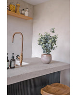 Een modern aanrecht met een bronzen kraan, zeepflessen, een houten kruk en de Kunstplant Eucalyptus 76 cm. Op de achtergrond zijn houten planken met potten en flessen gemonteerd op een lichtgrijze muur.