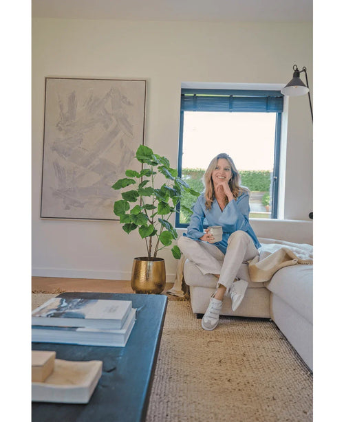 En kvinna sitter i en beige soffa i ett mysigt modernt vardagsrum, håller i en mugg och ler. Utrymmet kännetecknas av den konstgjorda Ficus-växten 120 cm i en guldkruka, abstrakta konstverk och solljus som skiner genom blå persienner.