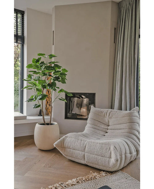 En mysig, beige fåtölj med struktur står bredvid den konstgjorda Ficusplantan 150 cm i ett modernt vardagsrum med ljusa trägolv, neutrala väggar, ett fönster och golvlånga grå gardiner.