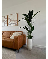 Een moderne woonkamer met een bruine leren bank, gestreepte kussens en de Kunstplant Heliconia 180 cm in een witte vaas. Daarboven hangt abstracte kunst aan een witte muur met een grijze vloer en een beige vloerkleed - ideaal voor kunstplant kopen inspiratie.