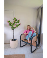 Een vrouw in een roze trui en blauwe spijkerbroek zit op blote voeten op een rotan stoel naast de Kunstplant Judas 150 cm, een kunstboom met roze bloemen, in een lichte, moderne kamer met witte muren en vloeren.