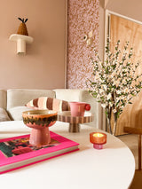 In een gezellige woonkamer staat een witte salontafel met een roze boek, schaal, sokkelschaal, kaars en de Kunstplant Kersenbloesem 120 cm crème. Op de achtergrond staan een beige bank met gestreepte kussens en roze behang met patroon.