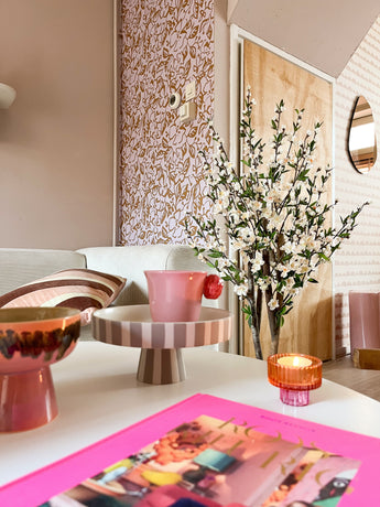 Een gezellige woonkamer met een roze en pastelkleurige inrichting is voorzien van een gestreept dienblad op voet, een roze kopje, een kaars, een schaal, een levendig tijdschrift en de Kunstplant Kersenbloesem 120 cm crème in een hoge vaas bij een gedessineerde muur en houten deur.