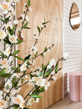 Een Kunstplant Kersenbloesem 120 cm crème met witte en lichtroze bloesems staat voor een houten deur. Daarachter zijn een roze kruk, spiegel en roze behang met patroon te zien.