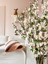 Een gezellige woonkamer met een witte bank, een gestreept roze en bruin kussen en de Kunstplant Kersenbloesem 120 cm crème bloeiende tak op de voorgrond. Een potplant rust op een wandplank tegen een lichtroze muur.